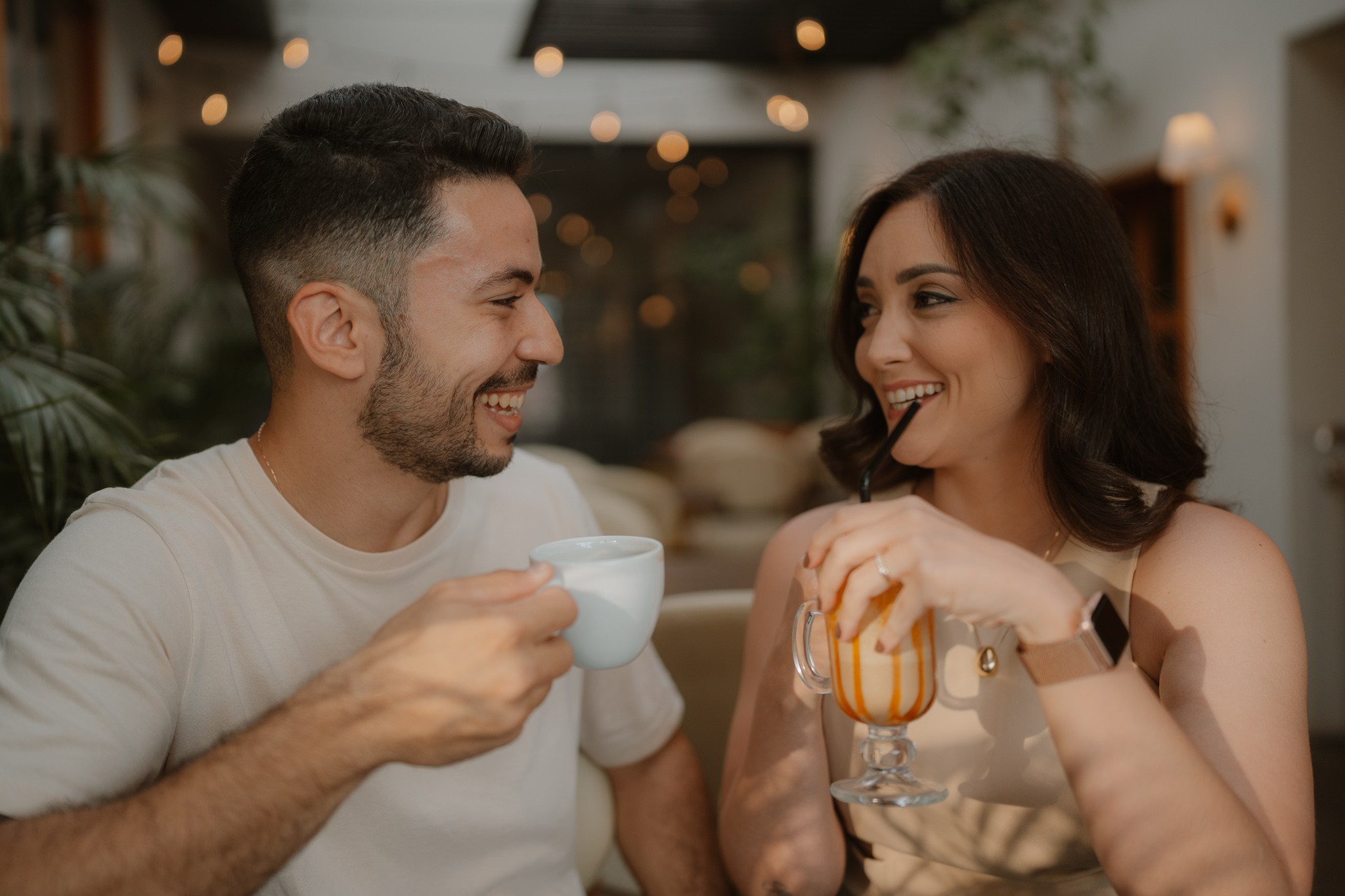 Casal sorrindo e brindando com café e bebida gelada durante ensaio lifestyle em cafeteria aconchegante em Presidente Prudente