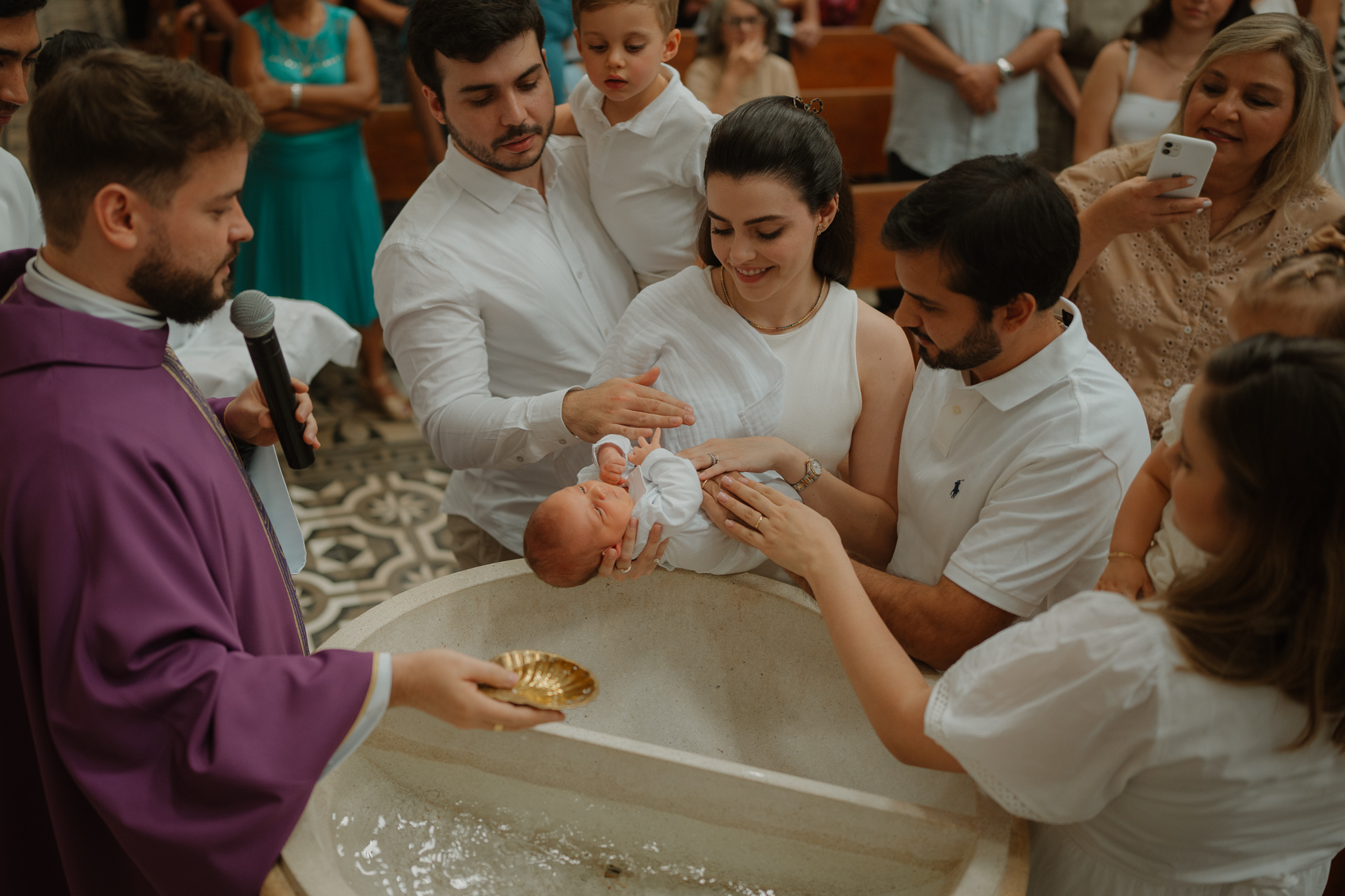 Foto do momento do batismo de um bebê em cerimônia católica com pais, padrinhos e sacerdote em Presidente Venceslau