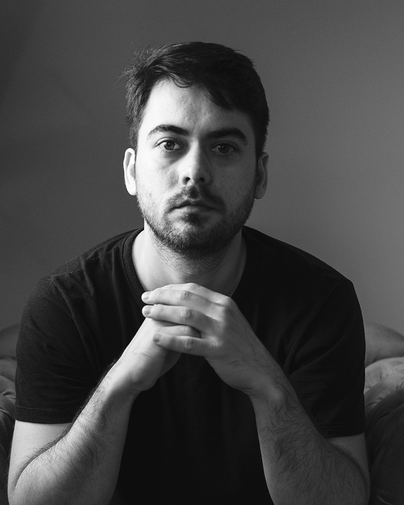 Retrato em preto e branco do fotógrafo Rafael Zolin, sentado e olhando diretamente para a câmera, com expressão séria e mãos entrelaçadas.