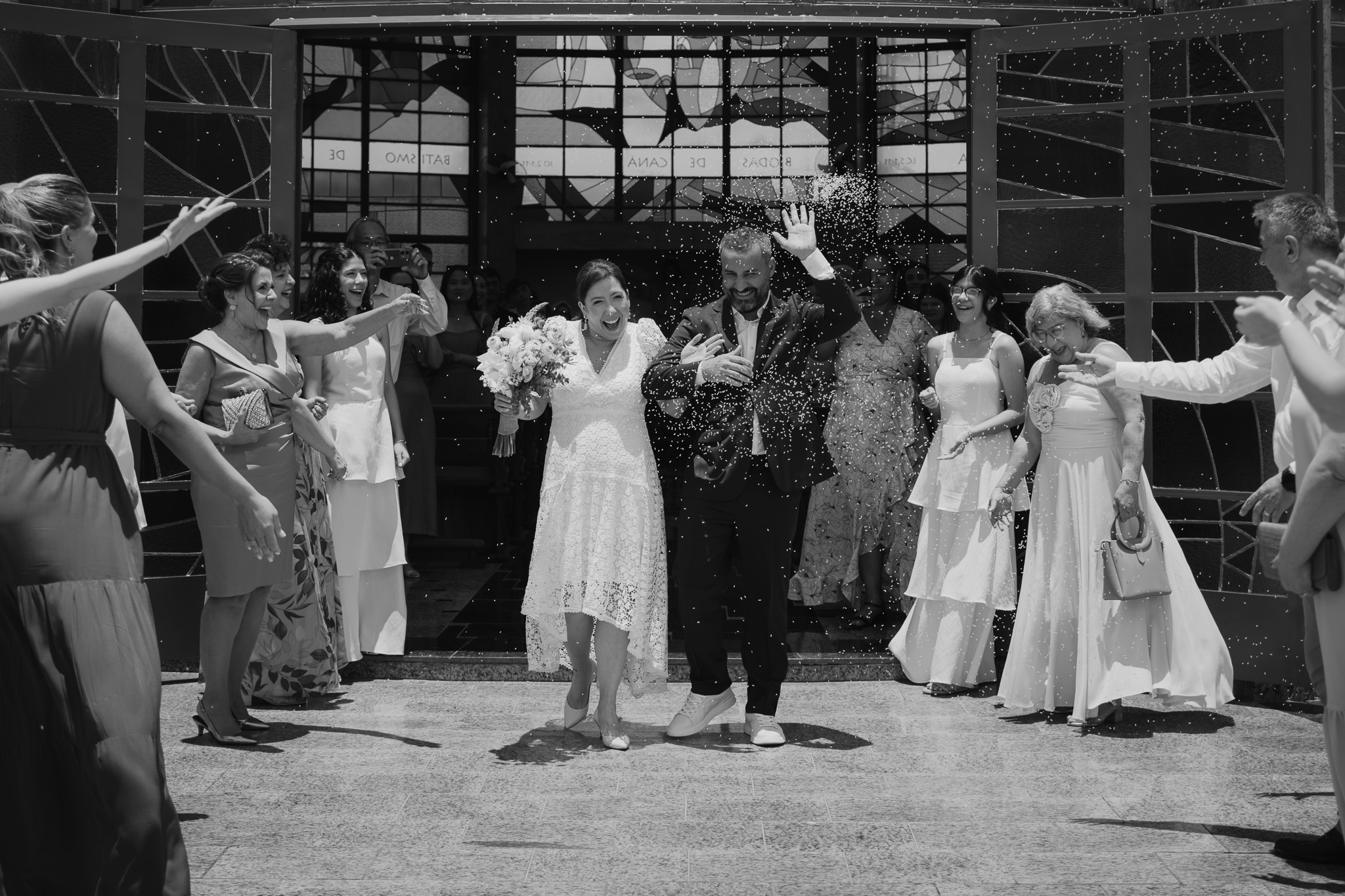 Noivos sorrindo ao sair da igreja sob chuva de arroz, rodeados por convidados, em foto documental de casamento em Presidente Prudente